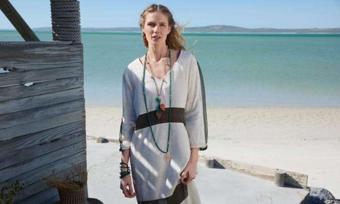 Mujer con túnica clara y collares largos de la colección India de Tamara Comolli, sobre fondo de playa